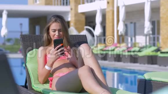 避暑胜地穿着泳衣手拿手机的漂亮女孩在昂贵的游泳池旁的休息室放松视频的预览图