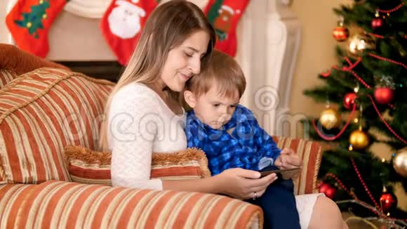 4k镜头快乐微笑的母亲和她的小儿子坐在圣诞树旁的扶手椅上看动画片视频的预览图
