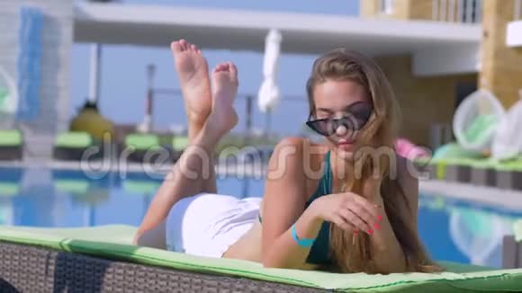 时尚迷人的女孩戴着太阳镜和泳衣在昂贵的度假胜地的泳池边休息在躺椅上视频的预览图
