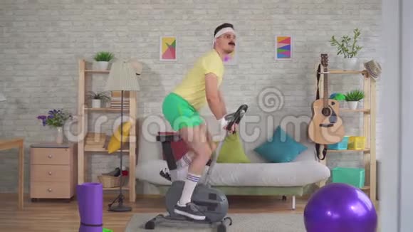 80年代留着胡子的滑稽运动员在公寓里骑着健身车视频的预览图