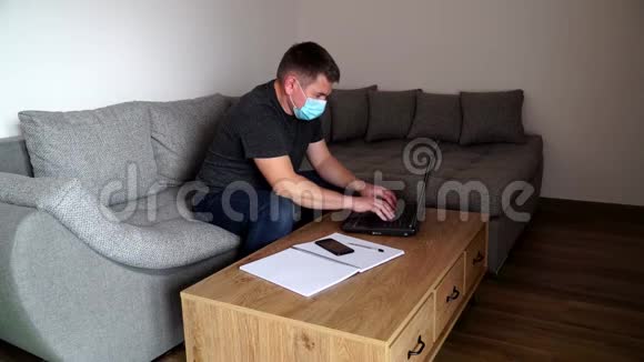 检疫冠状病毒COVID19戴着面具的年轻商人用智能手机说话坐着笔记本电脑办公室主任视频的预览图