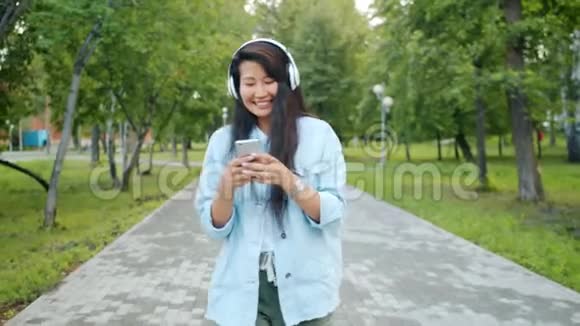 戴着耳机跳舞的快乐亚洲女孩用智能手机在公园里散步视频的预览图