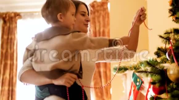 快乐微笑的母亲和她的孩子把彩色珠子和花环放在圣诞树上的4k视频家庭成员视频的预览图