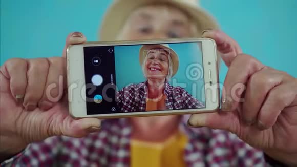 积极向上的老年微笑妇女拍摄自己的相机手机站在蓝色的背景通过观看视频的预览图