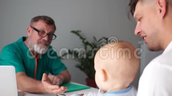 成熟的男儿科医生检查坐在年轻父亲手中的婴儿父亲日小儿科视频的预览图