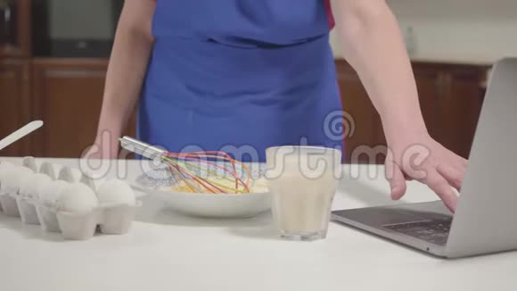 无法辨认的白种人成熟的女人在互联网上检查食谱把牛奶混合成面团家庭主妇退休视频的预览图