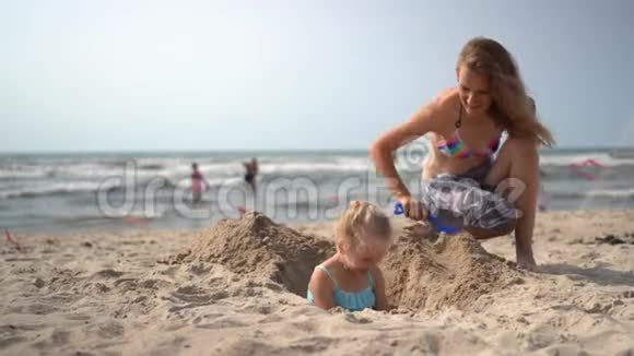 穿比基尼的母亲把她的金发小女孩埋在沙滩上摄像机运动视频的预览图