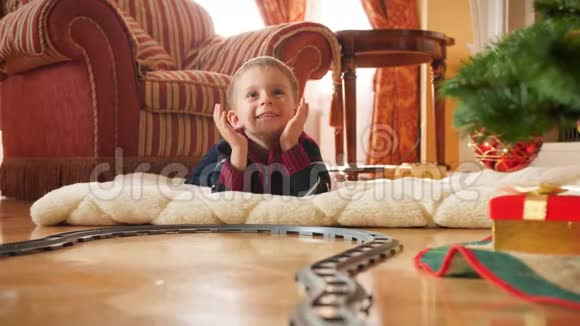 4k视频快乐微笑的小男孩躺在圣诞树旁边的地板上看着他的新玩具铁路视频的预览图