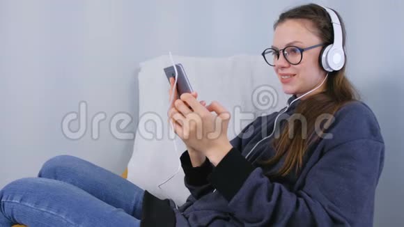 戴眼镜的女人在智能手机上用耳机听音乐坐在扶手椅上唱歌视频的预览图