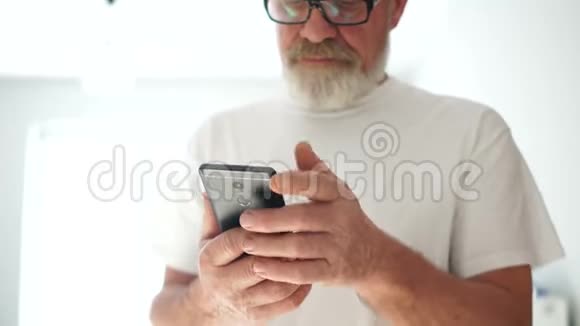 一位戴眼镜身穿白色T恤的灰胡子男子的近照正在使用智能手机微笑的成熟男人视频的预览图
