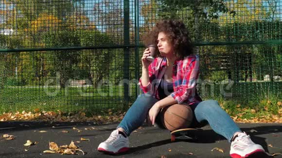 一个十几岁的小女孩坐在操场上的滑板上喝着纸杯里的咖啡拿着篮球视频的预览图