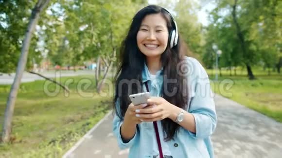 戴着耳机跳舞的亚洲女人带着智能手机在公园散步视频的预览图