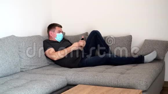一个戴着医用口罩的被隔离的冠状病毒携带者累了躺在沙发上打电话看社交节目视频的预览图
