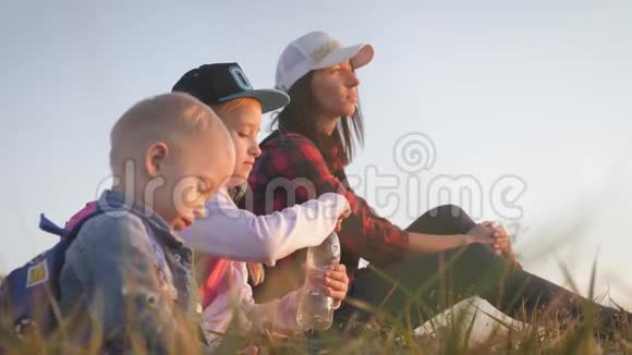 母亲和两个背包的孩子坐在山上的小径上他们喝水在旅途中放松视频的预览图
