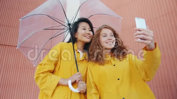 快乐女孩朋友们穿着雨衣撑着雨伞在户外自拍视频的预览图