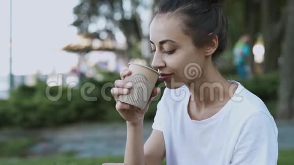 穿着白色t恤的漂亮年轻女子夏天晚上坐在公园的户外喝着纸杯里的咖啡视频的预览图