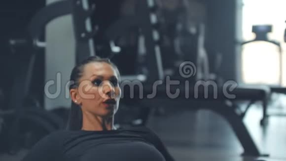 一个充满活力的女孩正在健身房做腹肌训练在一个跑步的男人的背景下在一个大窗户上做腹肌训练视频的预览图