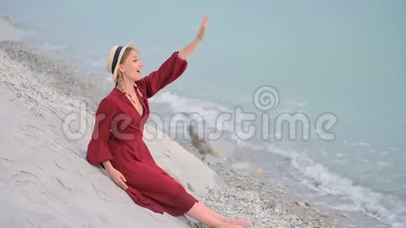 一个快乐的微笑女孩穿着红色的夏装和草帽坐在岩石海岸上看着大海的地平线他是视频的预览图