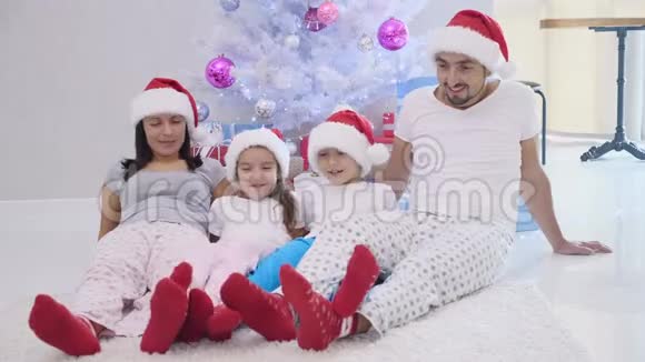 快乐的父亲母亲兄弟姐妹准备过圣诞节坐在杉树附近的地毯上戴着圣诞帽视频的预览图