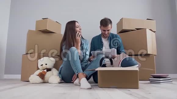 漂亮快乐的女孩和她英俊的男朋友坐在地板上打电话电话一边看报纸视频的预览图
