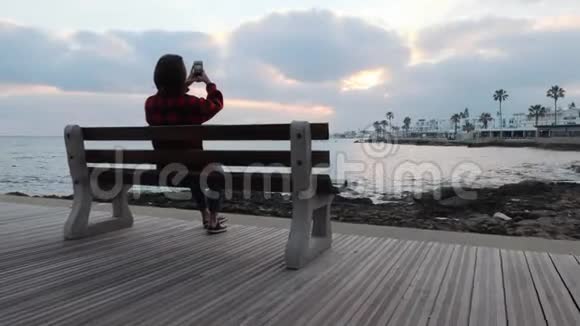年轻女子坐在长凳上拍海和日落的剪影女性在长廊拍照剪影视频的预览图