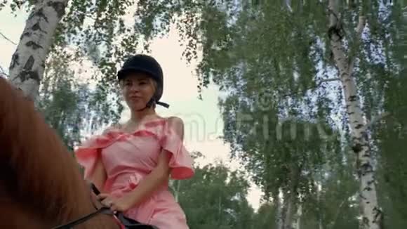 年轻迷人的女孩戴着保护头盔和粉红色的连衣裙坐在一匹棕色的马背上背景是树木和天空底部视频的预览图