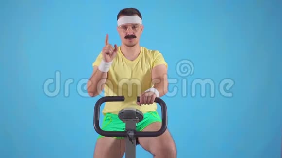 一位80岁留胡子的年轻滑稽男子的画像显示一只手指坐在运动自行车上视频的预览图