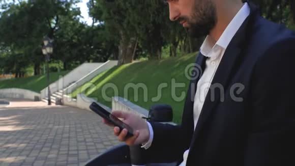 有胡子的年轻商人拿着一部手机靠近他的耳朵坐在公园的长凳上穿着经典西装的胡子男商务视频的预览图