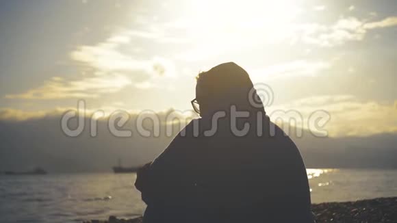 年轻女子在海滩上看日落或日出一个戴着眼镜和浴衣的女孩坐在海滩上视频的预览图