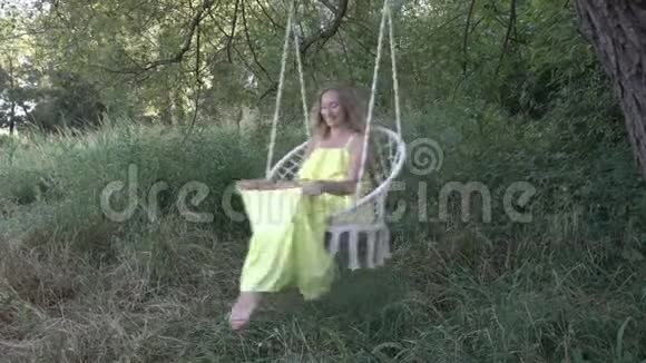 性感的金发女孩穿着黄色的裙子在户外的秋千上摇摆她微笑着光着脚摆姿势视频的预览图