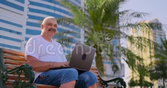 一位身穿白色t恤的老人坐在公园棕榈树间的长凳上看着笔记本电脑屏幕视频的预览图
