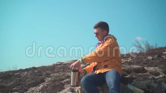 一个穿着黄色夹克蓝色牛仔裤和眼镜的年轻人坐在山上从热水瓶里倒了一杯茶视频的预览图