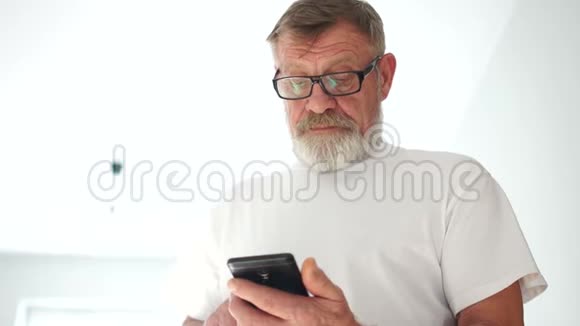 一个穿着白色T恤和眼镜的成熟的灰胡子男人在智能手机上拨通一个号码开始交谈老了视频的预览图