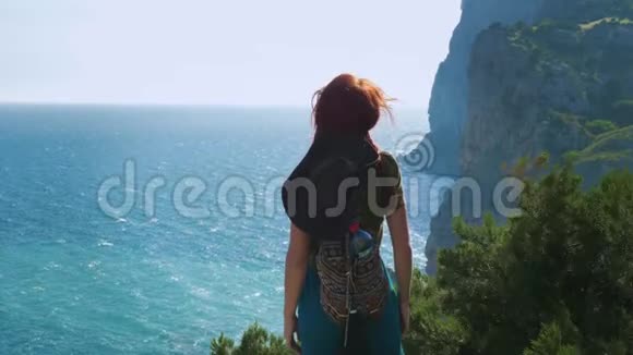 一个年轻的红发女孩旅行者戴着牛仔帽和背包站在山顶上双臂张开视频的预览图
