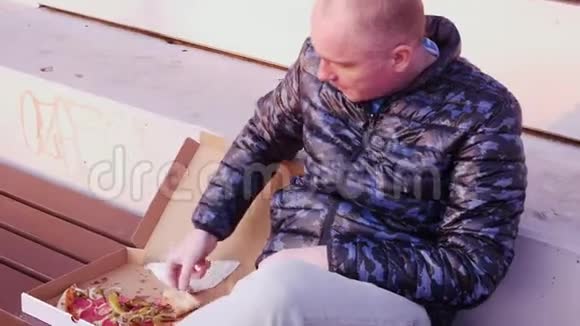 一个年轻的秃头坐在公园里从盒子里吃披萨视频的预览图