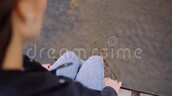 一个穿着皮夹克的女孩坐在木桥边晃着腿她的腿碰到了水喷水缓慢视频的预览图