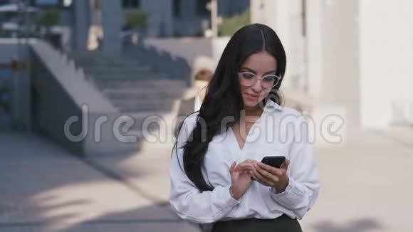 办公大楼附近使用智能手机的眼镜和白衬衫的成功商务女性画像视频的预览图