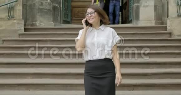 成熟的微笑积极的女人戴着眼镜穿着浅色衬衫走在楼梯上打着电话视频的预览图