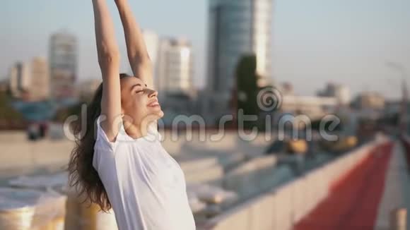 一位身穿白色t恤笑容满面的年轻漂亮女子举手站在城市背景的桥上视频的预览图