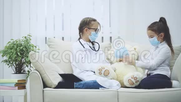 两个小女孩和白色泰迪熊坐在沙发上戴着医疗面具医生给熊一片药视频的预览图