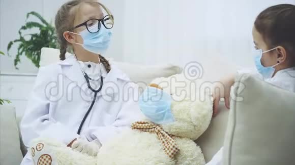 两个小女孩和白色泰迪熊坐在沙发上戴着医疗面具医生正在检查泰迪视频的预览图