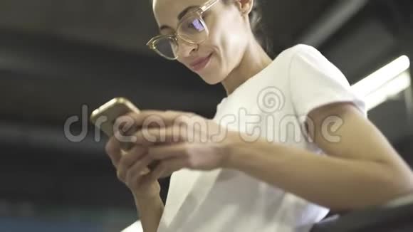 年轻的微笑时尚的美女穿着白色的t恤和透明的眼镜正在户外使用智能手机妇女视频的预览图