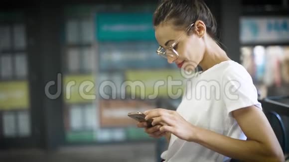 年轻的微笑时尚的美女穿着白色的t恤和透明的眼镜正在户外使用智能手机妇女视频的预览图