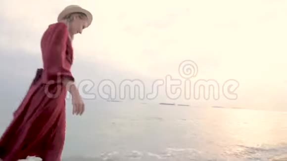 年轻迷人的微笑女孩穿着一件红色的浅色连衣裙和一顶草帽在风和日丽的天气里沿着海滨漫步视频的预览图