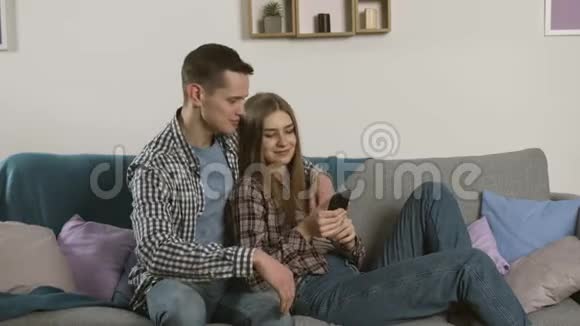 在沙发上用智能手机穿着格子衬衫微笑的情侣视频的预览图