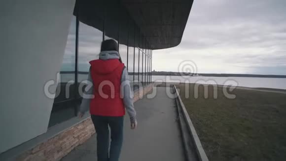 阴天穿红色无袖夹克的女孩走在大窗户旁边视频的预览图