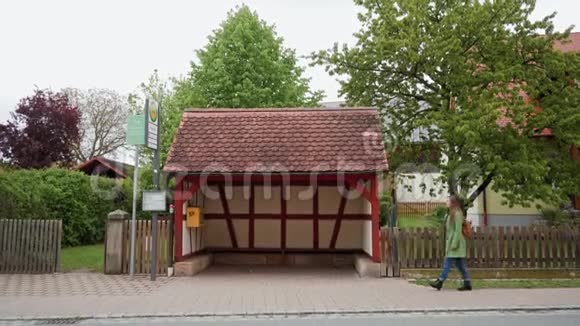 德国巴伐利亚Fachwerk式公共汽车站附近的一位穿着绿色外套和背包等待公共汽车的孤独女子视频的预览图