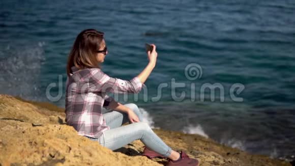 一个年轻的女人坐在海边的石头上拿着电话戴太阳镜的女孩海水飞溅视频的预览图