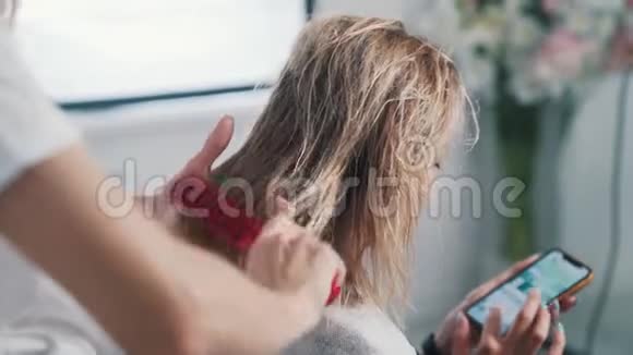 靠近理发师的手梳湿发的女人客户使用智能手机视频的预览图