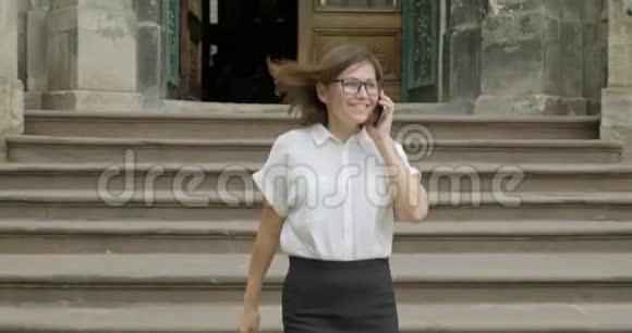 成熟的微笑积极的女人戴着眼镜穿着浅色衬衫走在楼梯上打着电话视频的预览图